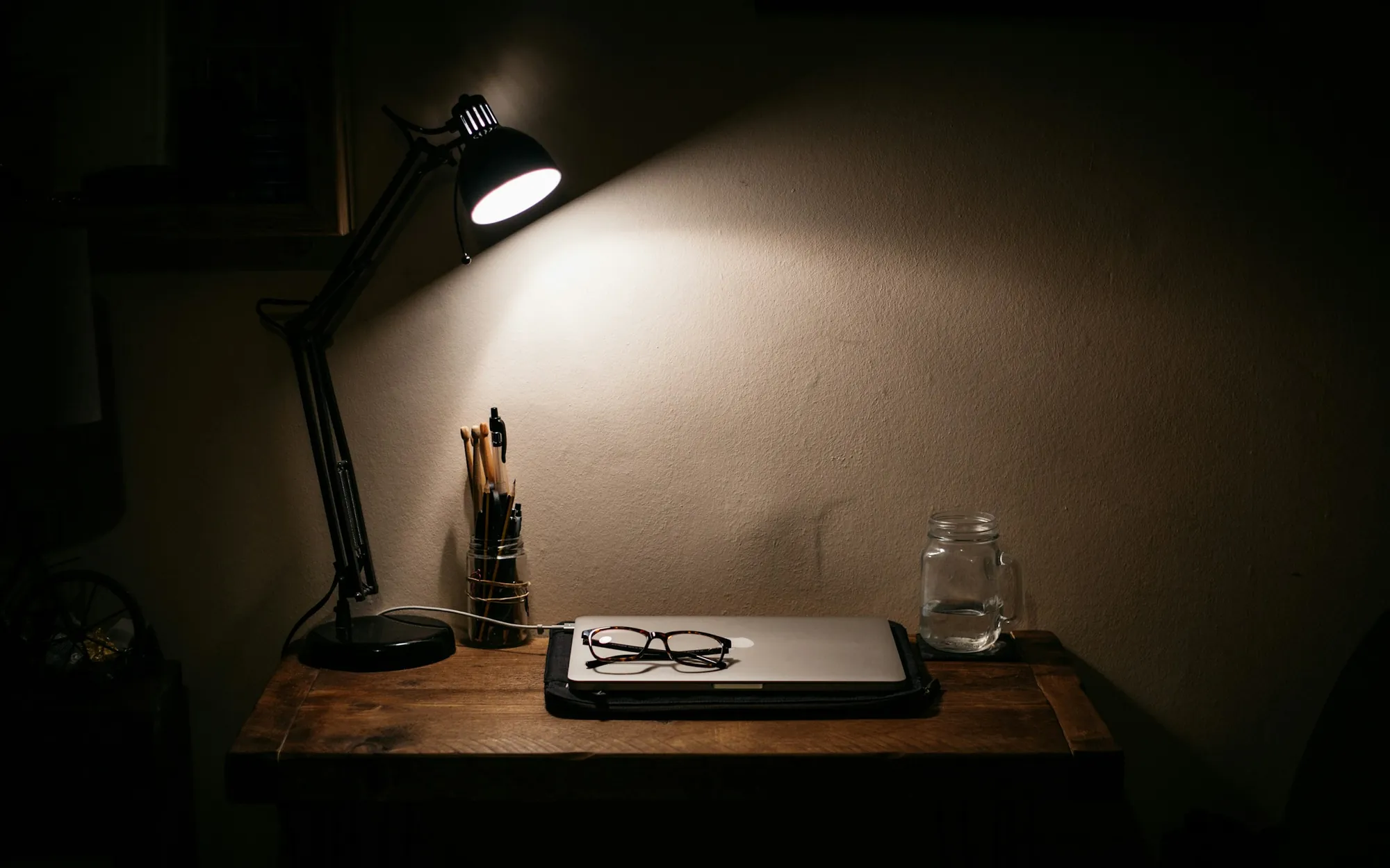 A desk lit by a single lamp in a dark room, with a closed laptop, reading glasses and a jar of pens.